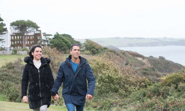 couple walking up along coastline with carlyon bay hotel the background