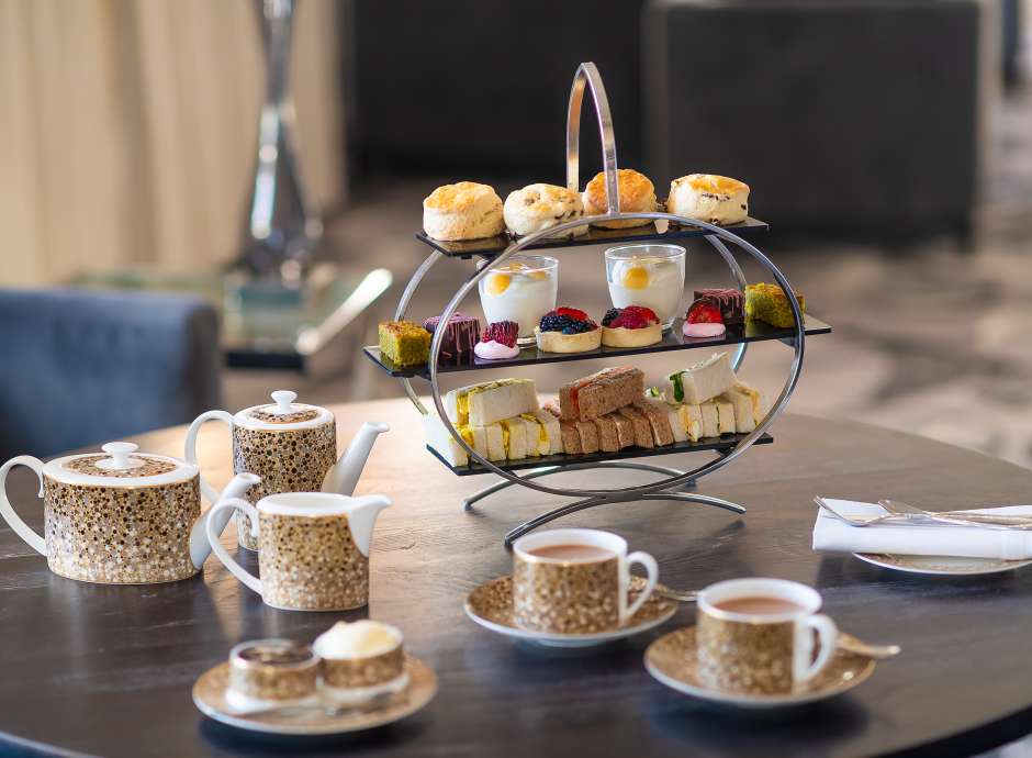 Afternoon tea rack on a table featuring scones, sandwiches and cakes with tea cups and tea pot