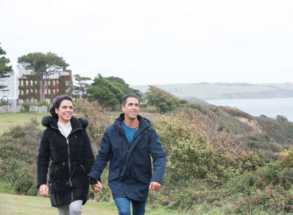 couple walking up along coastline with carlyon bay hotel the background